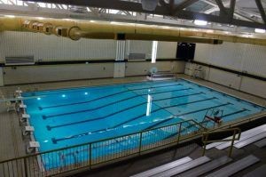 Indoor swimming pool with lane markers and diving boards, surrounded by spectator seating and metal railings.